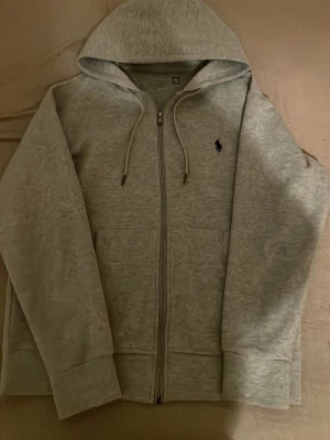 Grå zip hoodie från Polo Ralph Lauren - Snygg gråmelerad hoodie från Polo Ralph Lauren med dragkedja framtill och klassisk huva med dragsko. Ikoniska Polo-loggan broderad i mörkblått på bröstet. Mjuk bomullsblandning och två fickor framtill. Perfekt för en chill och stilren look.