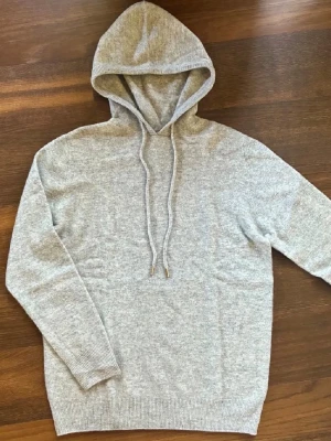 Merinoull hoodie - Mysig ljusgrå hoodie i stickad kvalitet med huva och långa ärmar. Hoodien har snörning med metalländar och ribbade muddar vid ärmslut och nederkant. Perfekt för en chill och avslappnad stil.