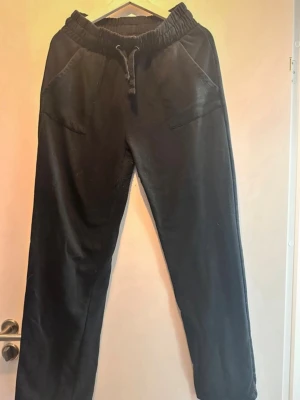 Svarta mjukisbyxor med snörning - Ett par svarta mjukisbyxor med dragsko i midjan och fickor både fram och bak. Byxorna har en relaxed passform och är perfekta för chill dagar. Tillverkade i mjukt material som känns skönt mot huden.