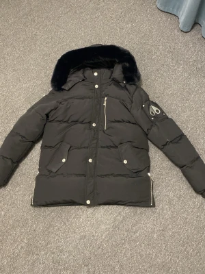 Svart dunjacka med fuskpäls från Moose Knuckles - Svart dunjacka från Moose Knuckles med avtagbar huva kantad av svart fuskpäls. Jackan har silverfärgade dragkedjor, tryckknappar och två stora fickor framtill. Logotyp på ärmen och quiltad design ger en cool och modern look.