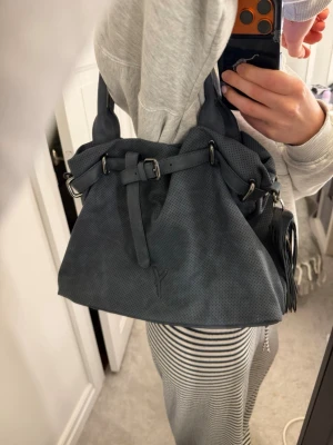 Mörkblå handväska  - Väskan har dubbla handtag, dekorativt bälte framtill och en tofsdetalj på sidan. Perfekt för dig som vill ha en stilren och trendig accessoar. Många fack