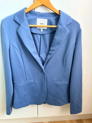 Blå kavaj från ICHI - Stilren blå kavaj från ICHI med klassisk krage och en knapp framtill. Kavajen har två fickor och är sydd i ett mjukt material som ger en bekväm känsla. Perfekt för att lyfta vilken outfit som helst. Storlek M, inga teckan på användning!