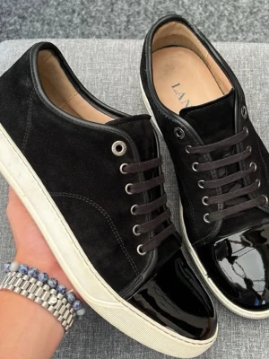 Svarta sneakers från Lanvin - Snygga svarta sneakers från Lanvin med ovandel i mocka och lackad tå. Skorna har vita sulor, svarta skosnören och metallfärgade öljetter. Perfekta för dig som vill ha en stilren och trendig look.