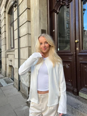 Vit design by si cardigan - Säljer en stilren vit kofta med en enkel knapp framtill. Koftan är tunn och har en avslappnad passform med långa ärmar. Perfekt att slänga över ett linne eller t-shirt för en clean look. Materialet ser ut att vara mjukt och lätt, troligen bomull eller viskos. Slutsåld på hemsidan, köpt för 600
