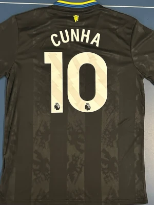 Manchester United Fotbollströja Cunha #10  - Säljer en svart Manchester United matchtröja med gula detaljer och Cunha #10 på ryggen. Tröjan har gula ränder på axlarna, klubbmärke och Snapdragon-logga på bröstet samt Premier League-märke på siffrorna. Storlek M. Helt ny.