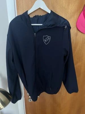 Svart DIF vindjacka med huva - Snygg svart vindjacka från DIF med huva och dragkedja framtill. Jackan har klubbens logga tryckt på bröstet och elastiska muddar vid ärmsluten. Perfekt för dig som vill visa ditt stöd för laget på ett stilrent sätt.