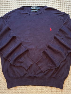 Mörkblå stickad tröja Polo Ralph Lauren i 100% pima bomull - Mörkblå stickad tröja från Polo Ralph Lauren med klassisk röd broderad logga på bröstet. Tröjan har rund halsringning, långa ärmar och ribbade muddar vid ärmslut och nederkant. Perfekt för lager på lager och stilren look. 100% pima bomull. Mycket bra skick, inga defekter förutom lappen är lös i ett hörn (väldigt lätt lagning). Storlek L. Axelbredd: 51 cm Längd: 66 cm