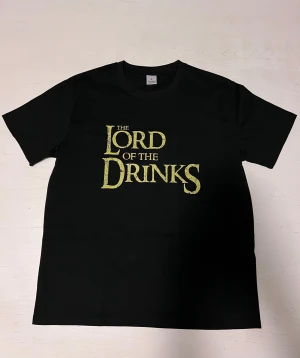 ”The Lord Of The Drinks” T-shirt - En svart t-shirt i storlek S med ett humoristiskt tryck som parodierar “Lord of the Rings”. Texten “The Lord of the Drinks” är tryckt i guldfärgad, sliten stil som ger en cool och unik look. Tröjan är i nytt skick och perfekt för någon som gillar roliga motiv eller temafester.