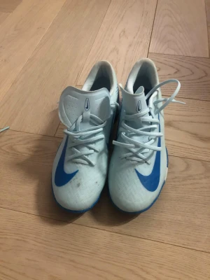 Nike vita och blå fotbollsskor - Snygga fotbollsskor från Nike i vitt med blå detaljer och klassisk swoosh på sidan. Skorna har snörning och är tillverkade i syntetmaterial för bra grepp och känsla på planen. Perfekta för dig som vill sticka ut med stil på fotbollsplanen.