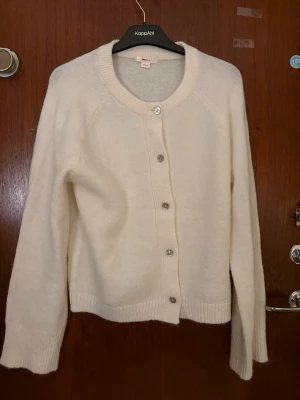 Vit kofta från KappAhl - Mysig vit kofta från KappAhl med rund halsringning och långa ärmar. Koftan har ribbade muddar och knäpps med silvriga knappar framtill. Perfekt att slänga över en topp för en enkel och clean look.