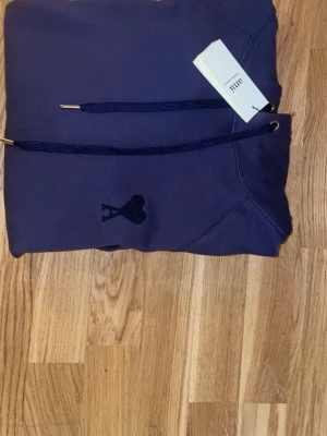 Mörkblå hoodie från Ami Paris - Snygg mörkblå hoodie från Ami Paris med klassisk huva och dragsko. På bröstet finns Ami Paris ikoniska hjärtemblem i matchande färg. Hoodien har känguruficka framtill och ribbade muddar vid ärmar och nederkant. Perfekt för en avslappnad och trendig look. 