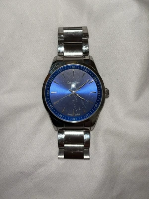Calvin Klein blå klocka i stål - Snygg klocka från Calvin Klein med blå urtavla och silverfärgat armband i rostfritt stål. Klockan har minimalistisk design med tydliga indexmarkeringar och klassisk rund form. Perfekt accessoar för dig som gillar stilrena detaljer. Ny pris: 1890:- 