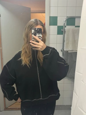 Svart oversized hoodie med kontrastsömmar - Svart oversized hoodie med dragkedja och vita kontrastsömmar. Hoodien har en avslappnad passform och långa ärmar. Perfekt för en chill och trendig look. Materialet ser ut att vara mjukt och bekvämt, troligen bomullsblandning.