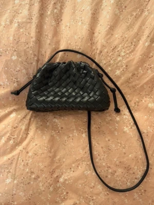 Svart flätad axelväska i skinn - Snygg svart axelväska med flätad design och justerbar axelrem. Väskan har en mjuk, rundad form och stängs med snörning upptill. Perfekt för dig som gillar stilrena accessoarer med en twist.