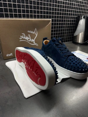Mörkblå Christian Louboutin sneakers - Säljer ett par mörkblå sneakers från Christian Louboutin med coola nitar på tån och sidorna. Skorna har snörning, vit sula med röd undersida och är tillverkade i mocka. Insidan är beige och de har en rund tå. Perfekt för dig som gillar statement pieces. Kan byta mot gats eller andra saker (helst skor) 