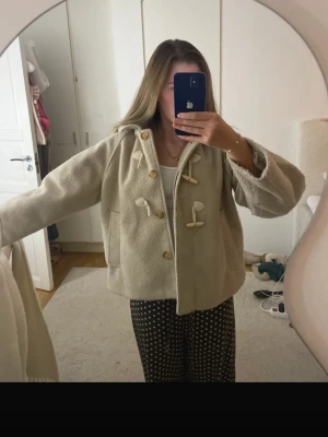 Beige kappa med träknappar - Snygg beige kappa med huva och träknappar i toggle-stil. Jackan har en boxig passform och är tillverkad i ett ull-liknande material. Perfekt för dig som gillar en stilren och mysig look.