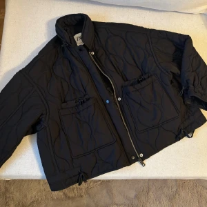 Svart quiltad pufferjacka från Zara - Snygg svart pufferjacka från Zara med quiltat mönster och två stora framfickor. Jackan har hög krage, dragkedja och tryckknappar framtill samt justerbar nederkant. Perfekt för kalla dagar och riktigt trendig look. Jackan har även en inbyggd luva 
