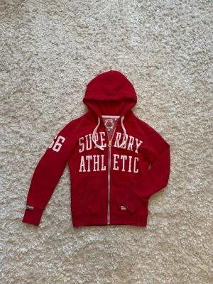 Superdry zip hoodie röd - Snygg hoodie med dragkedja från superdry, använd men i gott skick.  Mått: Längd: 61 Bröstvidd: 49 Ärm: 62
