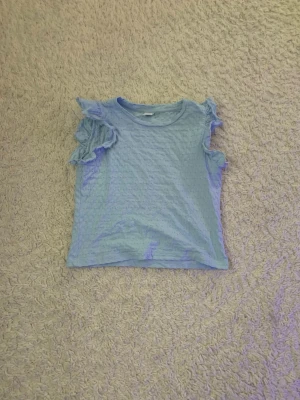 Ljusblå topp med volang från Lindex - Supersöt ljusblå topp från Lindex med volangdetaljer på axlarna. Tillverkad i mjuk bomull med diskret strukturmönster i tyget. Perfekt för dig som vill ha en söt och bekväm topp till vardagslooken.