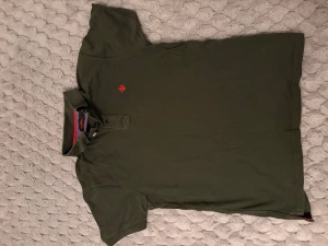 Mörkgrön pikétröja från Morris - Klassisk mörkgrön pikétröja från Morris med röd broderad logga på bröstet. Tröjan har korta ärmar, krage och knappar vid halsen. Perfekt för en avslappnad men ändå stilren look.