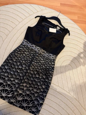 Svart och silvermönstrad kort klänning - Snygg svart ärmlös klänning med silvrigt mönster på kjolen. Överdelen är enfärgad och kjolen har ett elegant grafiskt mönster i silver. Perfekt för dig som vill sticka ut med en stilren look. Klänningen har en figurnära passform och rund halsringning.