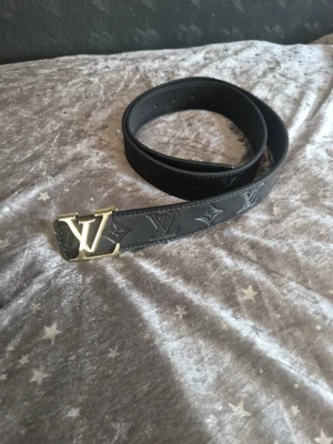 Louis Vuitton svart läderbälte - Snyggt svart bälte från Louis Vuitton i äkta läder med klassiskt LV-monogram präglat längs hela bältet. Spännet är i guldmetall med den ikoniska LV-loggan. Perfekt accessoar för att lyfta vilken outfit som helst.