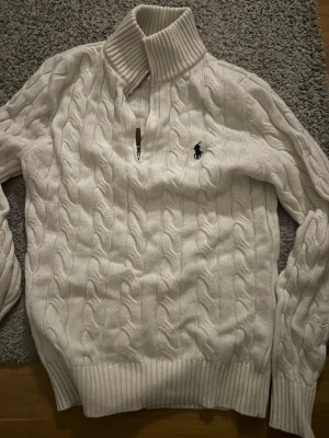 Vit kabelstickad tröja Polo Ralph Lauren - Vit kabelstickad tröja från Polo Ralph Lauren med halv dragkedja och hög ribbad krage. Tröjan har det klassiska mörkblå logobroderiet på bröstet och ribbade muddar vid ärmslut och nederkant. Perfekt för en clean och stilren look.