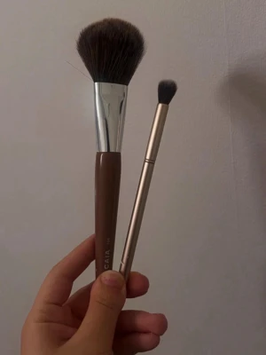 CAIA sminkborstar duo - Säljer två sminkborstar från CAIA – en större puderborste med brunt handtag och täta, mjuka borst samt en mindre borste med metallfärgat handtag och rundad topp. Perfekta för applicering av puder, highlighter eller ögonskugga. Båda borstar har syntetiska borst. Skriv för intresse! 