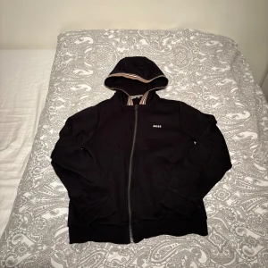 Svart hoodie från BOSS med dragkedja JR - Svart hoodie från BOSS med dragkedja framtill och liten vit logga på bröstet. Huvan har ett snyggt randigt innerfoder och tröjan har ribbade muddar vid ärmslut och nederkant. Perfekt för en stilren och avslappnad look. Finns dock ett litet hål bredvid loggan som inte syns jättemycket.