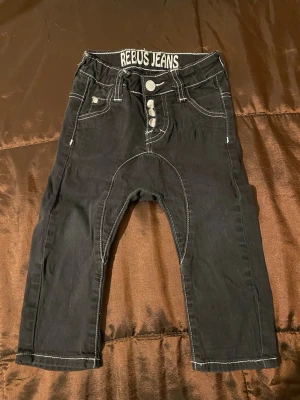 Svarta Rebus Jeans med vita sömmar - Säljer ett par tuffa svarta barn  jeans från Rebus Jeans med tydliga vita kontrastsömmar och  knappar i gylfen. Jeansen har låg gren och klassisk femficksdesign. 