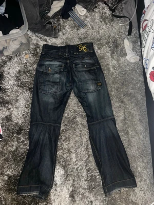 Mörkblå jeans med detaljerad sömnad - Snygga mörkblå jeans från G-Star Raw med coola sömdetaljer över benen och flera fickor bak. Jeansen har en rak passform och är tillverkade i klassiskt denimtyg. Perfekta för dig som gillar streetwear och vill sticka ut lite extra.