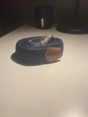 Mörkblå flätat bälte med läderdetaljer - Snyggt mörkblått flätat bälte med bruna läderdetaljer och silverspänne. Längden på den är 110 cm och är i nytt skick. 