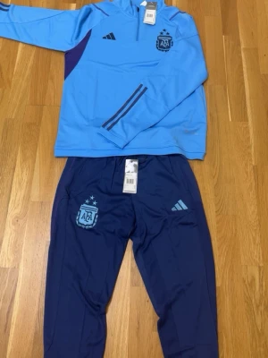 Argentina träningsset från Adidas - Snyggt träningsset från Adidas med Argentina AFA-logga. Överdel i ljusblått med dragkedja och mörkblå detaljer, samt matchande mörkblå byxor med ljusblå loggor. Tillverkat i funktionsmaterial som andas, perfekt för fotbollsträning.