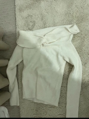 Off shoulder vit stickad tröja - Trendig off shoulder stickad tröja i vit färg med ribbad struktur. Tröjan har långa ärmar och en bred krage som kan stylas över axlarna för en cool look. Perfekt för dig som vill ha en enkel men snygg stickad topp.