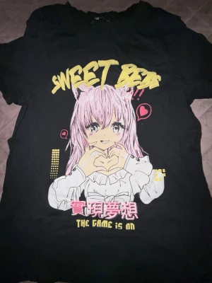 Svart anime t-shirt med tryck - Cool svart t-shirt med färgglatt anime-tryck framtill. Motivet visar en tjej med rosa hår och kattöron, samt texten 'SWEET DREAM', japanska tecken och 'THE GAME IS ON'. Perfekt för dig som gillar anime och streetstyle.