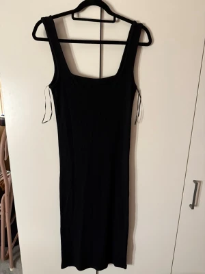 Svart ribbad långklänning - En svart, ribbad långklänning med breda axelband och fyrkantig ringning. Klänningen har en kroppsnära passform och är ärmlös, perfekt för en stilren och enkel look. Materialet känns stretchigt och bekvämt.