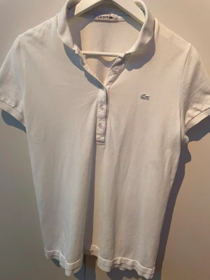 Vit pikétröja från Lacoste - Klassisk vit pikétröja från Lacoste med korta ärmar och krage. Tröjan har knappar framtill och den ikoniska krokodilloggan på bröstet. Tillverkad i mjuk bomull som känns skön mot huden. Perfekt till jeans för en clean look.