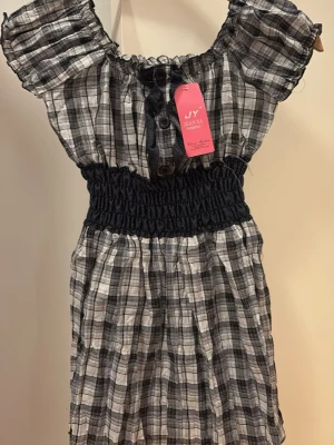 Rutig offshoulder blus - Trendig offshoulder blus i svartvitt rutigt mönster. Blusen har korta puffärmar, knappar framtill och en bred smock i midjan som ger en snygg siluett. Tillverkad i ett lätt och luftigt tyg, perfekt för en avslappnad look. Står ingen storlek men passar nog folk som bär xs-m