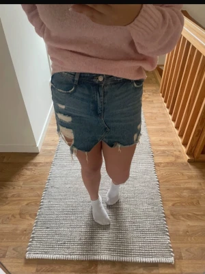 Blå jeanskjol med slitningar - Trendig blå jeanskjol med rå kant och slitna detaljer framtill och baktill. Kjolen har klassisk femficksdesign och knappgylf. 