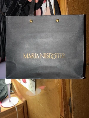 Svart påse från Maria Nilsdotter - Stilren svart påse från Maria Nilsdotter med guldfärgad logga på framsidan. Väskan har två handtag och en enkel, fyrkantig form som ger en klassisk look. Perfekt för dig som gillar minimalistisk design.