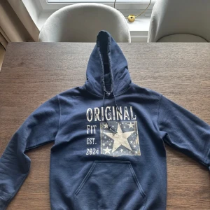 Marinblå hoodie med stjärntryck - Säljer en marinblå hoodie ifrån SCNDI Tröjan har huva med snörning och en klassisk ficka framtill. Hoddien är skön och kommer passa perfekt till våren och sommarkvällar.