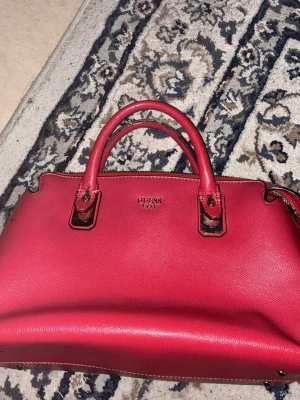 Röd handväska från Guess - Snygg röd handväska från Guess med två stadiga handtag och guldfärgade detaljer. Väskan har en klassisk, strukturerad form och stängs med dragkedja. Perfekt för dig som vill ha en färgklick till din outfit.