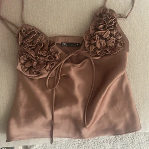 Brun satinlinne med volangdetaljer Zara - Snyggt brunt satinlinne från Zara med tunna axelband och dekorativa volangdetaljer över bysten. Linnet har en glansig finish och knytband framtill som ger en trendig look. Perfekt för att lyfta vilken outfit som helst.