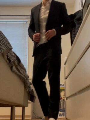 Mörkblå kostym Riley - Snygg mörkblå kostym med en enkelknäppt kavaj och matchade mörkblåa kostymbyxor till. Med en vit skjorta under kavajen. Jag är 183 lång och väger 73-75 kg och allt sitter bra på mig men på gränsen till att de är för litet! Vill du inte köpa hela kostymen utan till exempel bara kavajen osv är det bara att skriva så kommer vi fram till ett pris:) Skickar även med en kostymgaderob på köpet!