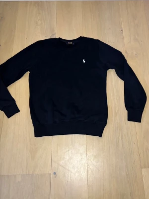 Mörkblå sweatshirt från Polo Ralph Lauren - Klassisk mörkblå sweatshirt från Polo Ralph Lauren med rund halsringning och den ikoniska vita broderade loggan på bröstet. Tröjan har ribbade muddar vid ärmslut och nederkant, perfekt för en clean och stilren look.