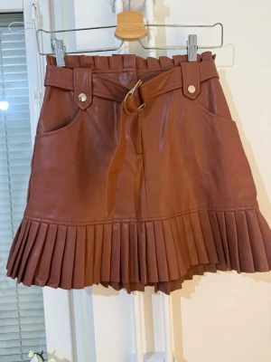 Brun plisserad skinnkjol med bälte  - Kort brun kjol i skinn med plisserad nederkant och tillhörande bälte i midjan. Kjolen har fickor fram, bred linning och dekorativa knappar. Perfekt för en trendig och edgy stil. 