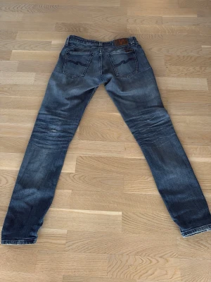 Blå slim fit jeans från Nudie Jeans - Säljer ett par blå slim fit jeans från Nudie Jeans med slitningar och snygg tvätt. Klassisk femficksmodell med läderpatch bak i midjan och kontrastsömmar på bakfickorna. Perfekta för dig som gillar en smalare passform och streetig look.