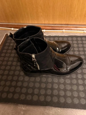 Svarta stövletter med dragkedja - Snygga svarta stövletter i blankt skinn och mocka med spetsig tå och dragkedja på sidan. Snygg quiltad detalj vid hälen och låg klack. Perfekta för dig som vill ha en edgy och stilren look.