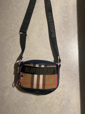 Rutig axelväska från Burberry - Säljer en klassisk axelväska från Burberry med det ikoniska rutiga mönstret i beige, svart, vitt och rött. Väskan har en justerbar axelrem med Burberry-logga och dragkedja framtill. Perfekt för dig som vill ha en stilren och trendig accessoar.