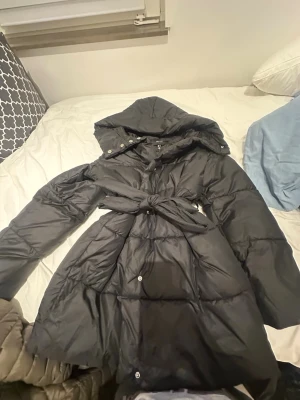 Svart lång dunjacka med huva och bälte - Snygg svart dunjacka med lång passform, stor huva och knytbälte i midjan. Jackan har quiltad design, långa ärmar och tryckknappar framtill. Perfekt för kalla vinterdagar och ger en stilren look.